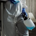 Person in PPE suit and gloves with disinfectant and cloth cleaning indoors.