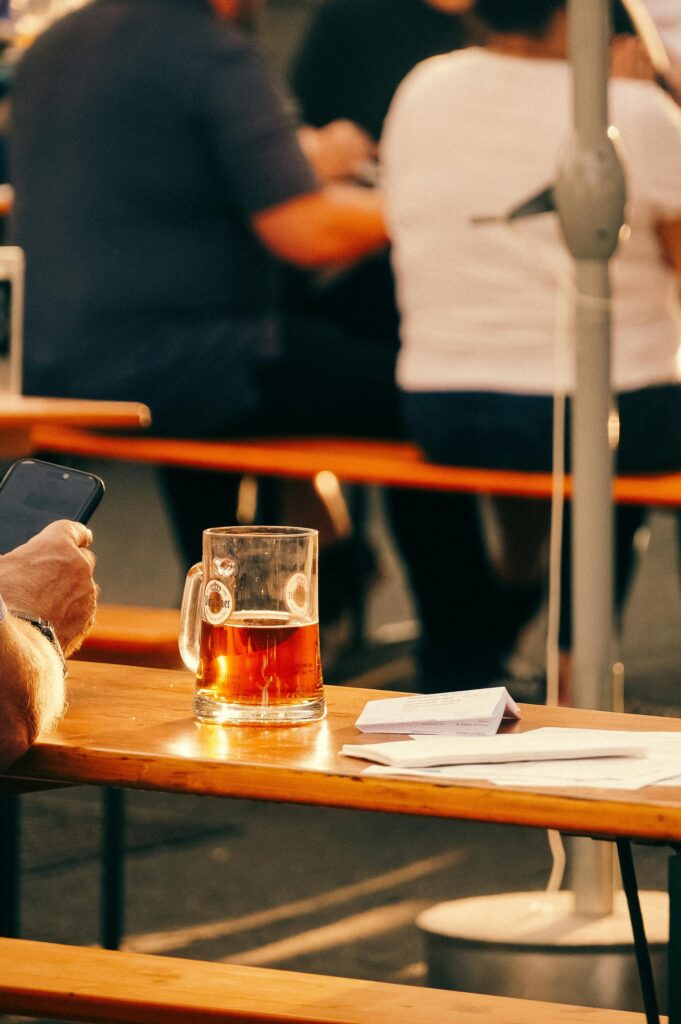 A sunlit beer garden with people enjoying drinks and using phones on a summer day.